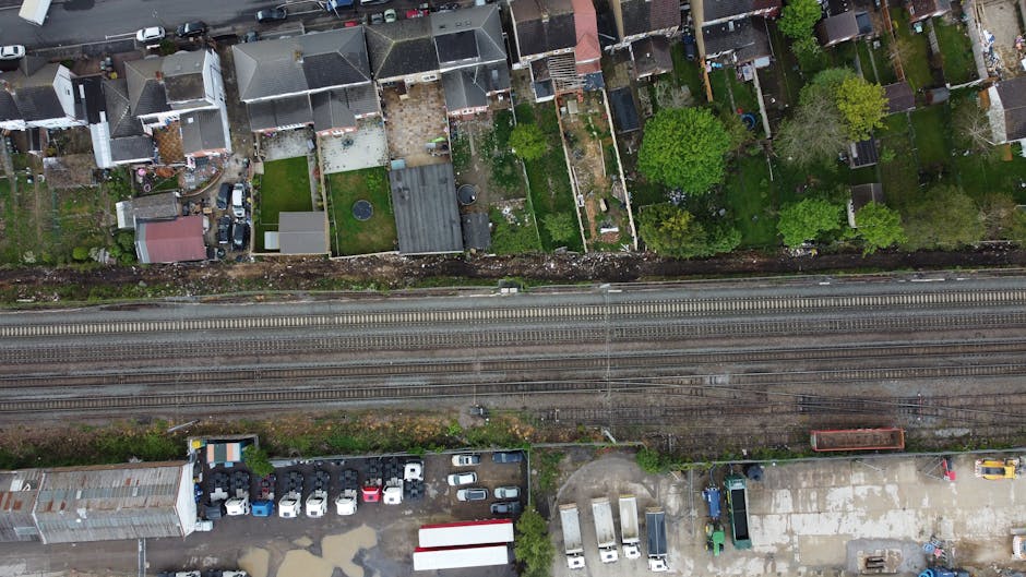 An aerial view of a residential area showing a row of houses with garden backyards featuring various plants, trees, and lawn areas. Some gardens have neatly trimmed hedges and flowerbeds, while others